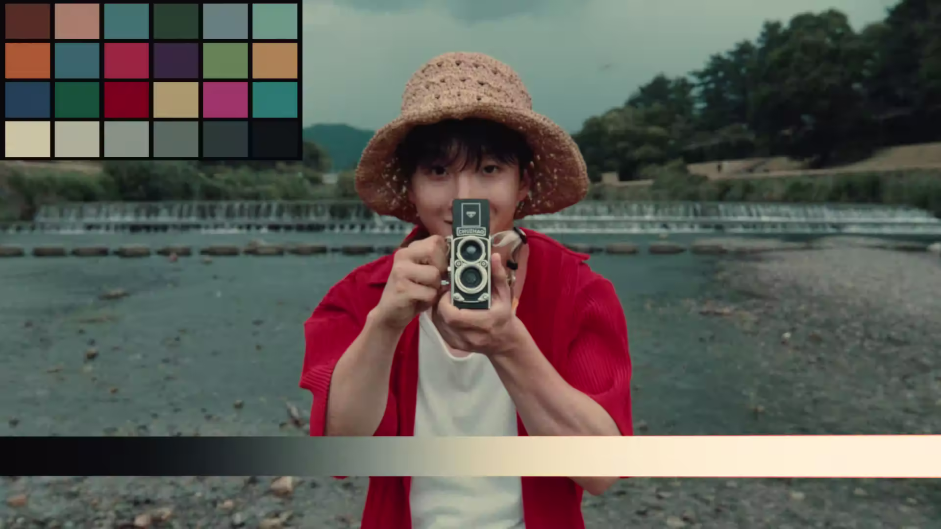 Person wearing a straw hat and red shirt holding a vintage camera pointed at the viewer, standing by a shallow river with a small waterfall and green trees in the background.