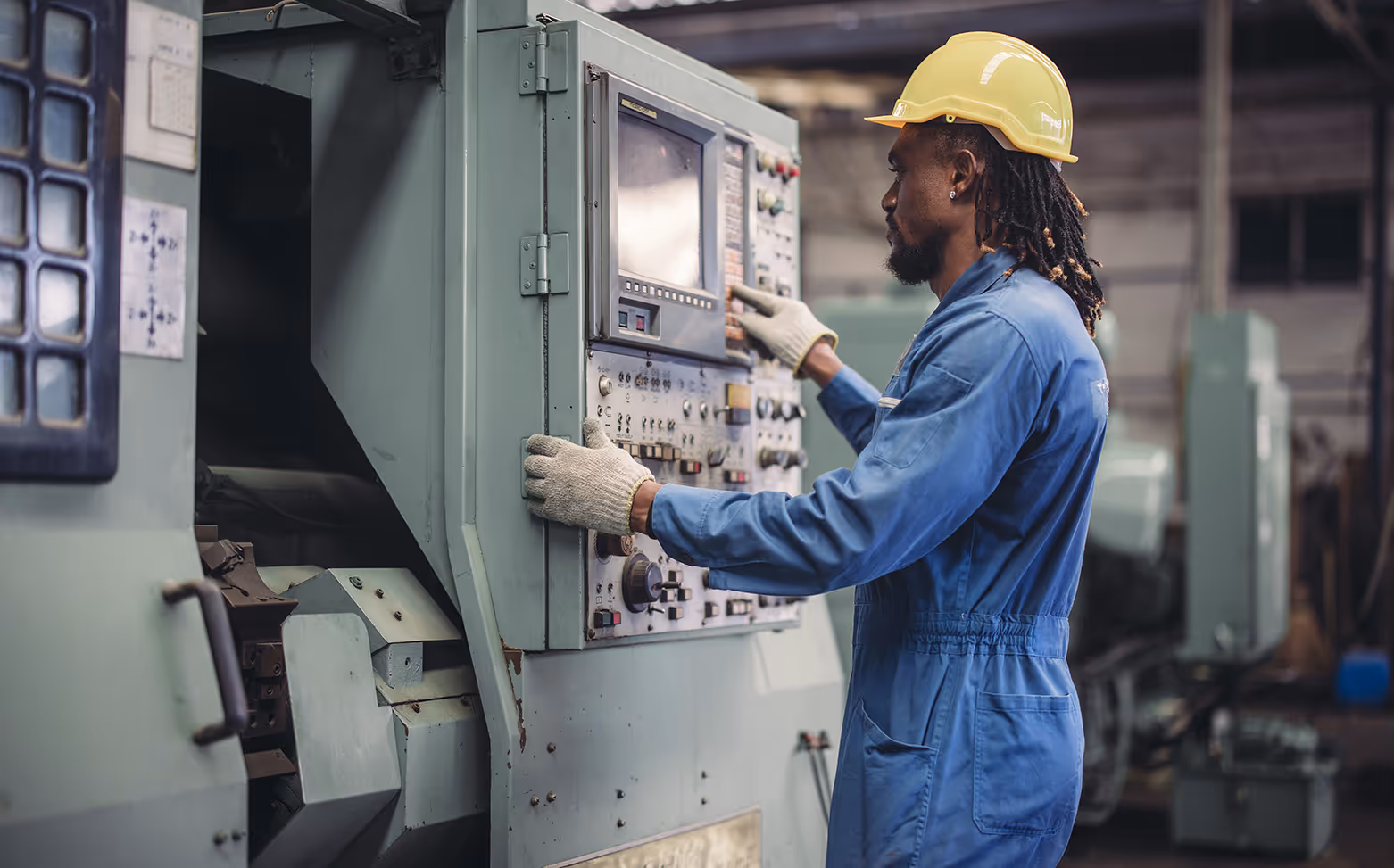Industrial worker in blue coveralls and yellow hard hat operating a control panel on a large machine.