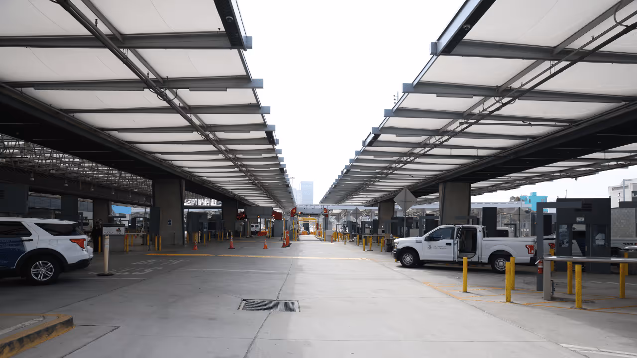 View down a wide concrete vehicle inspection or checkpoint area with overhead canopies and vehicles parked along the sides.