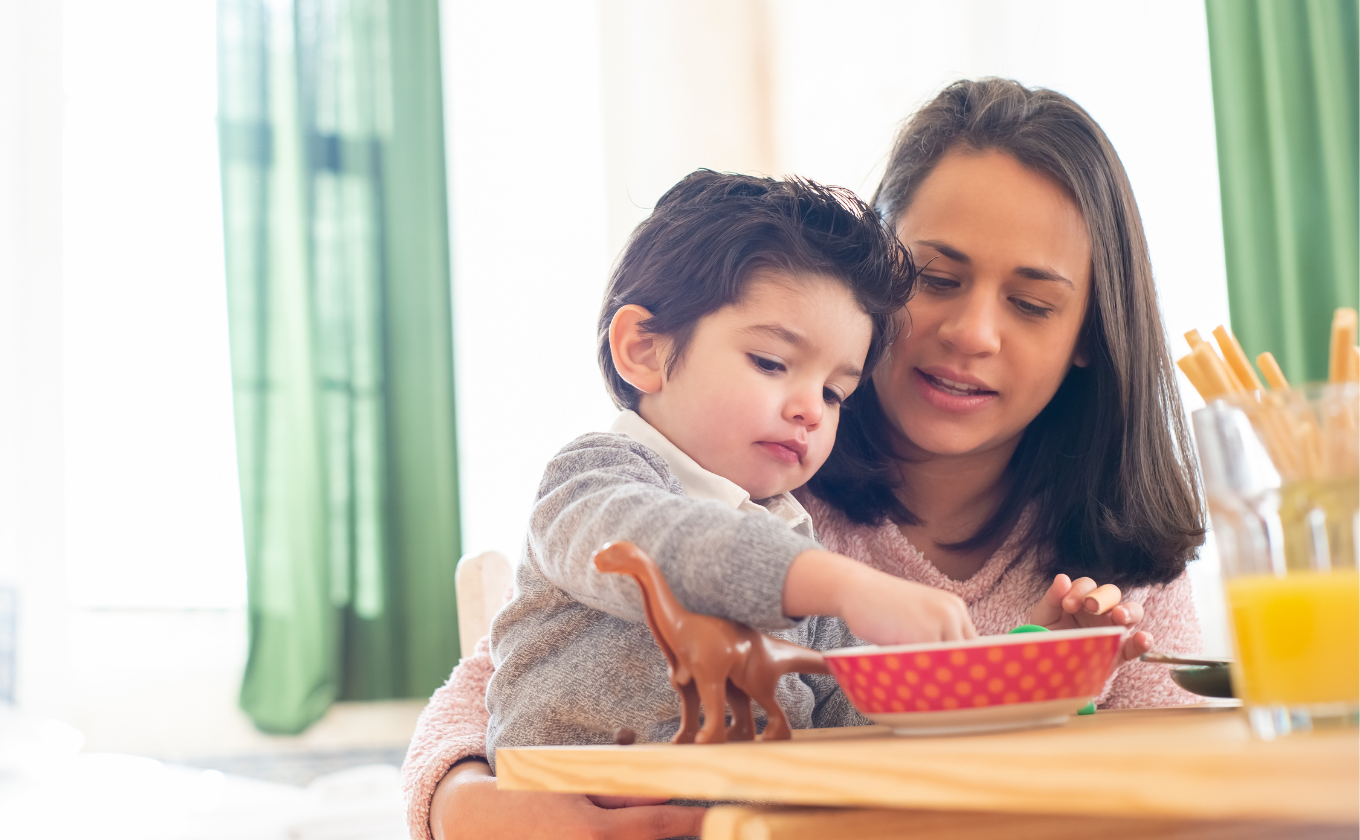 Woman helping a young boy eat from a bowl at a table with a toy dinosaur and juice nearby.