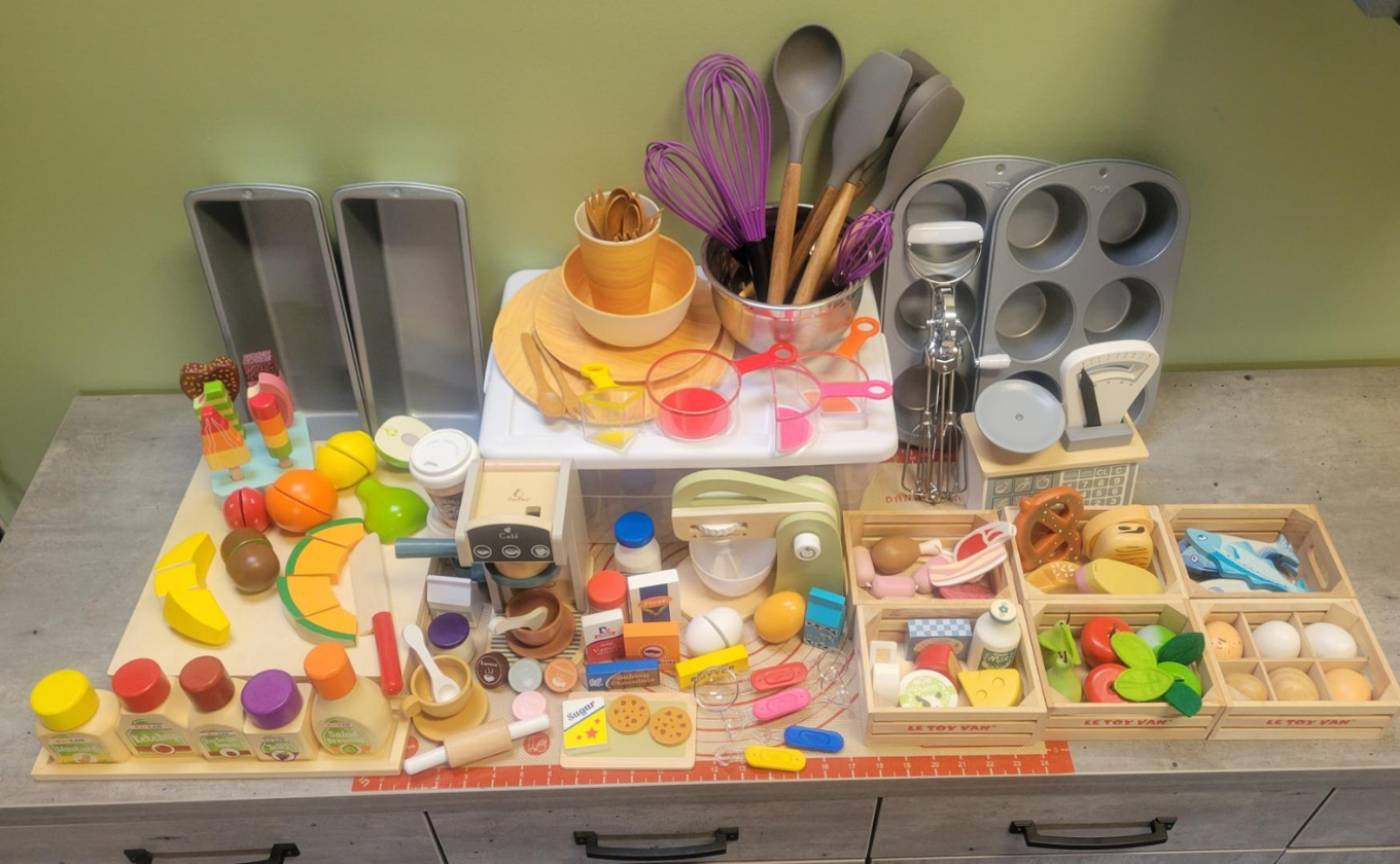 Toy baking set with utensils, measuring cups, mixing tools, pretend food, and baking trays on a table.