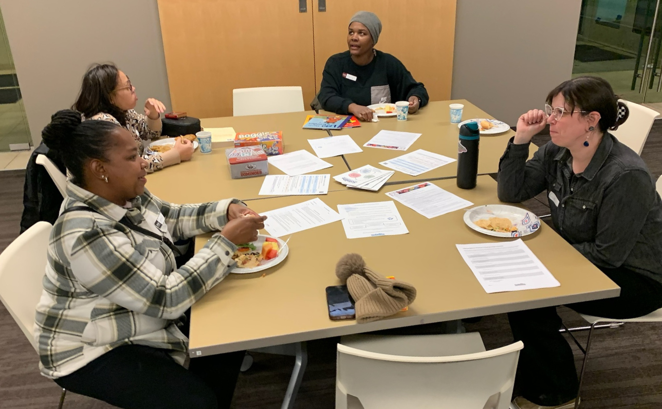 Four people eating and talking around a table with papers, snacks, and drinks in a meeting room.