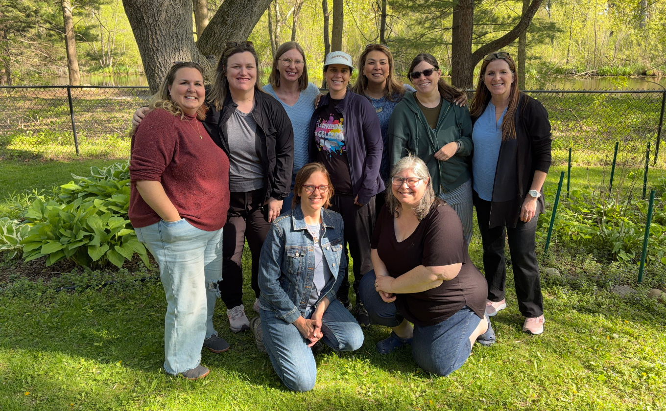 Group of nine women smiling outdoors on grass with trees and a fence in the background.