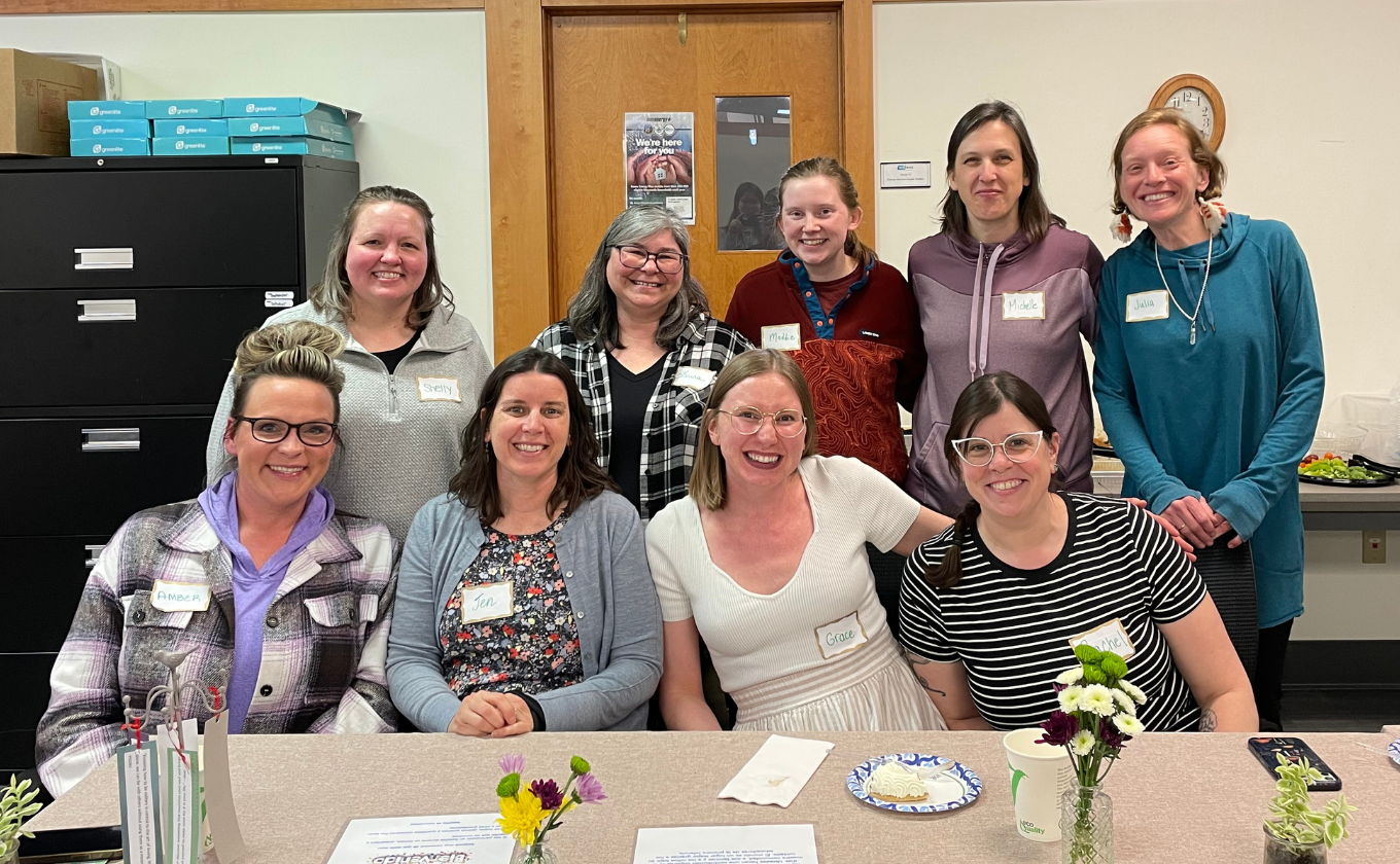 Group of ten smiling women wearing name tags seated and standing around a table with flowers and papers.