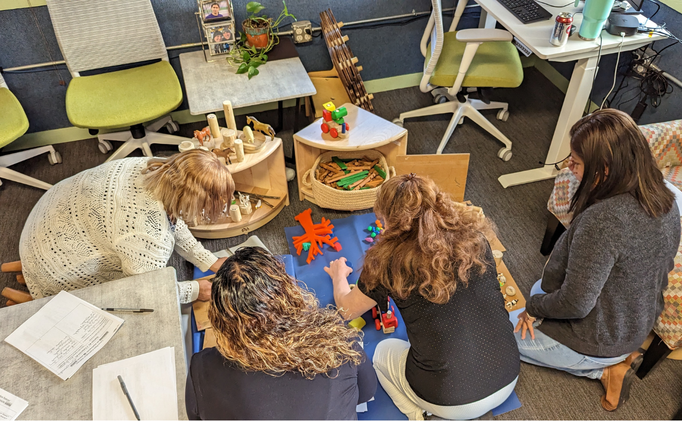 Four women kneeling on floor around wooden blocks and toys in an office setting with chairs and tables.