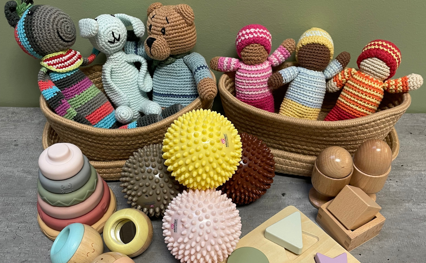 Crocheted dolls in baskets with colorful stacking rings, spiked sensory balls, and wooden toys on table.