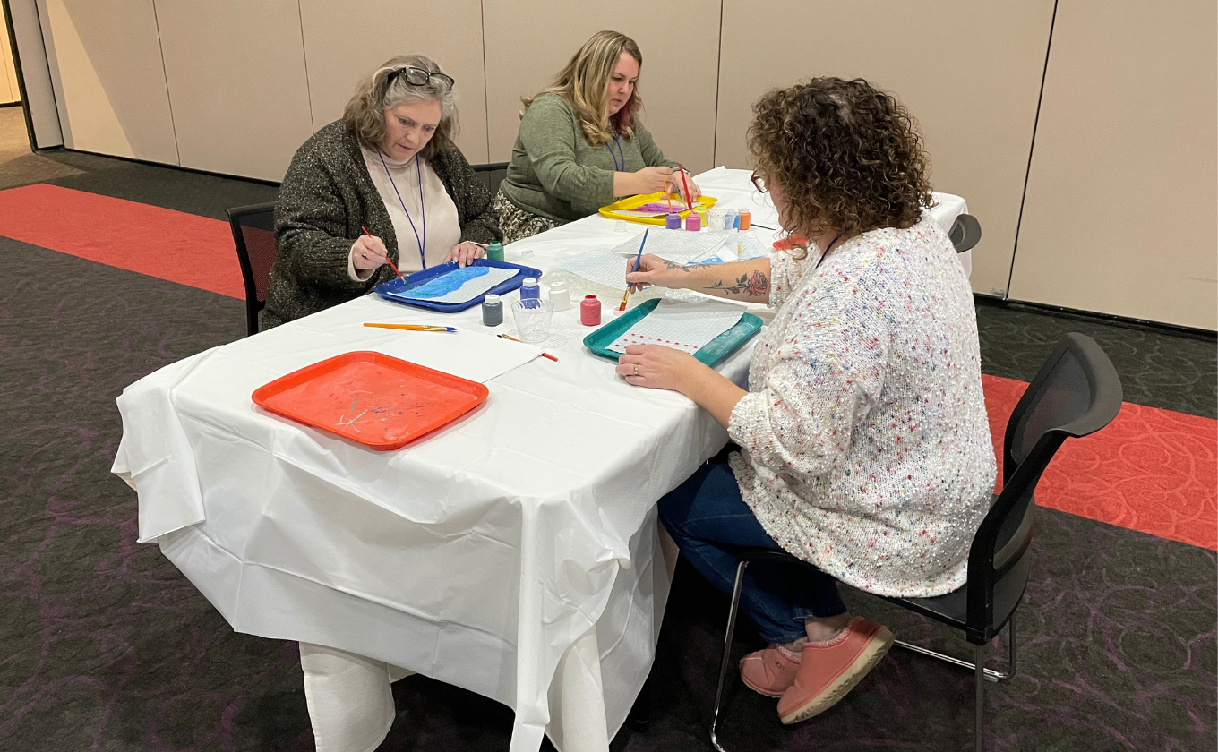 Three women seated at a table covered with a white cloth, painting on plastic trays.