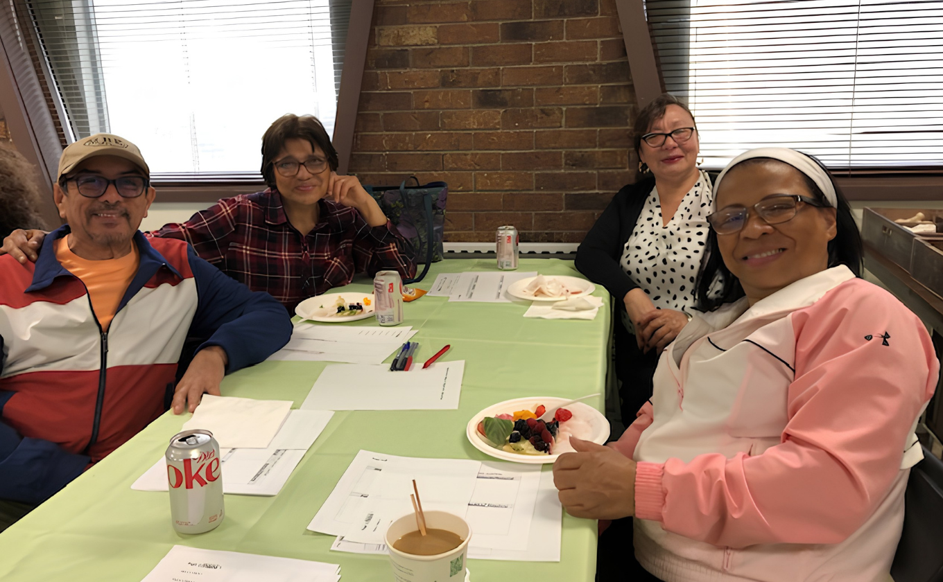 Four people sitting at a table with papers, drinks, and plates of fruit engaged in conversation.