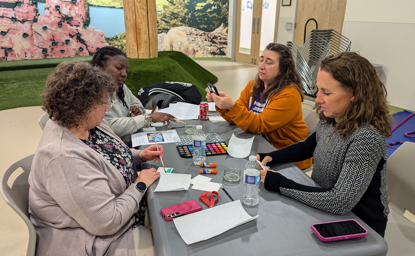 Four women seated around a table painting and crafting with watercolors, glue sticks, and scissors.