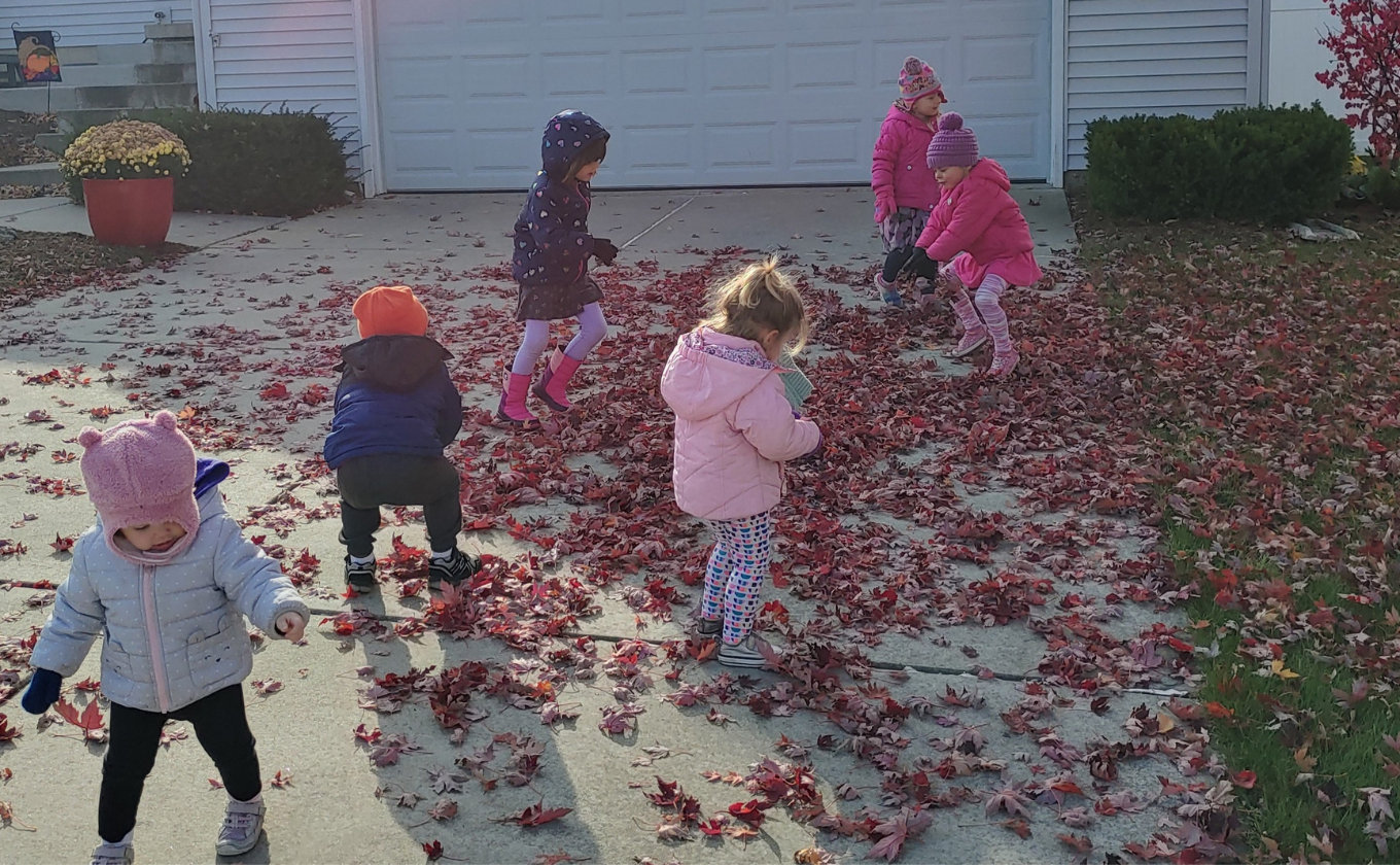 Five young children dressed in winter clothes playing among fallen autumn leaves on a driveway.