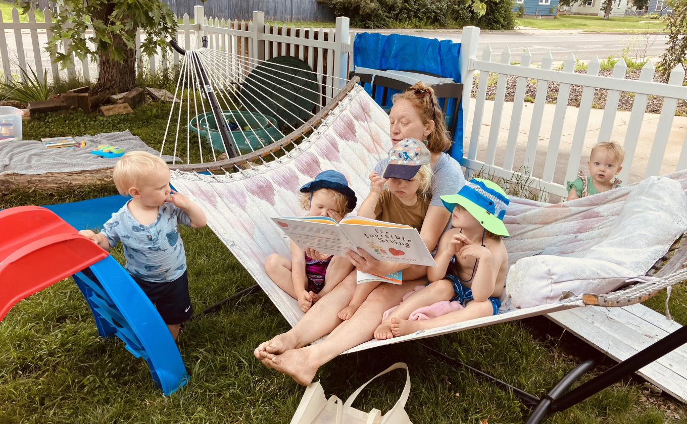 Adult reads book to four young children, three sitting on hammock and one standing nearby in garden.