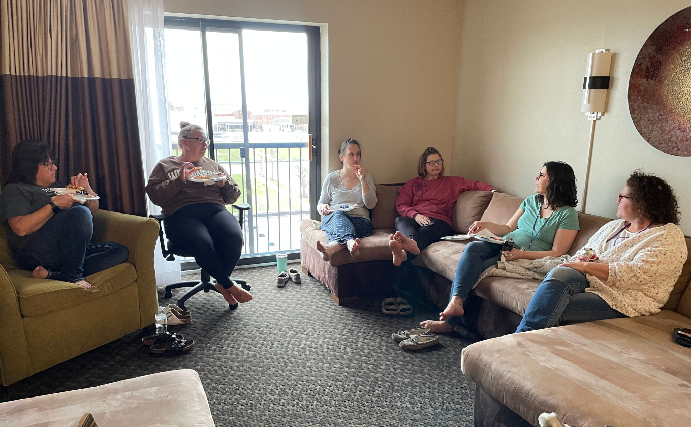 Six women sitting in a living room chatting and eating while barefoot.