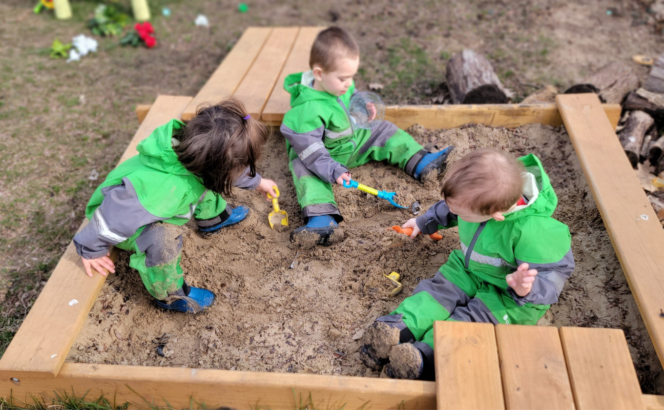 Three toddlers in green rain suits playing with shovels in a sandbox outdoors.