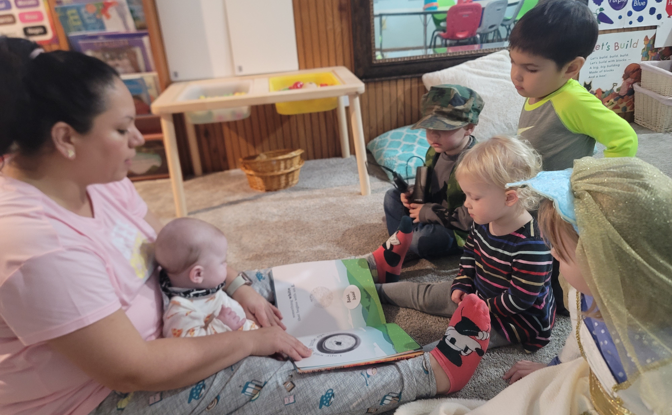 Woman reading a book to a baby and a group of young children sitting on a carpeted floor.