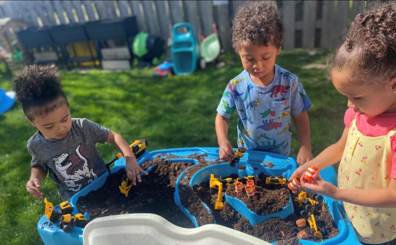Three children playing with toy construction vehicles and dirt in a blue outdoor sensory table.