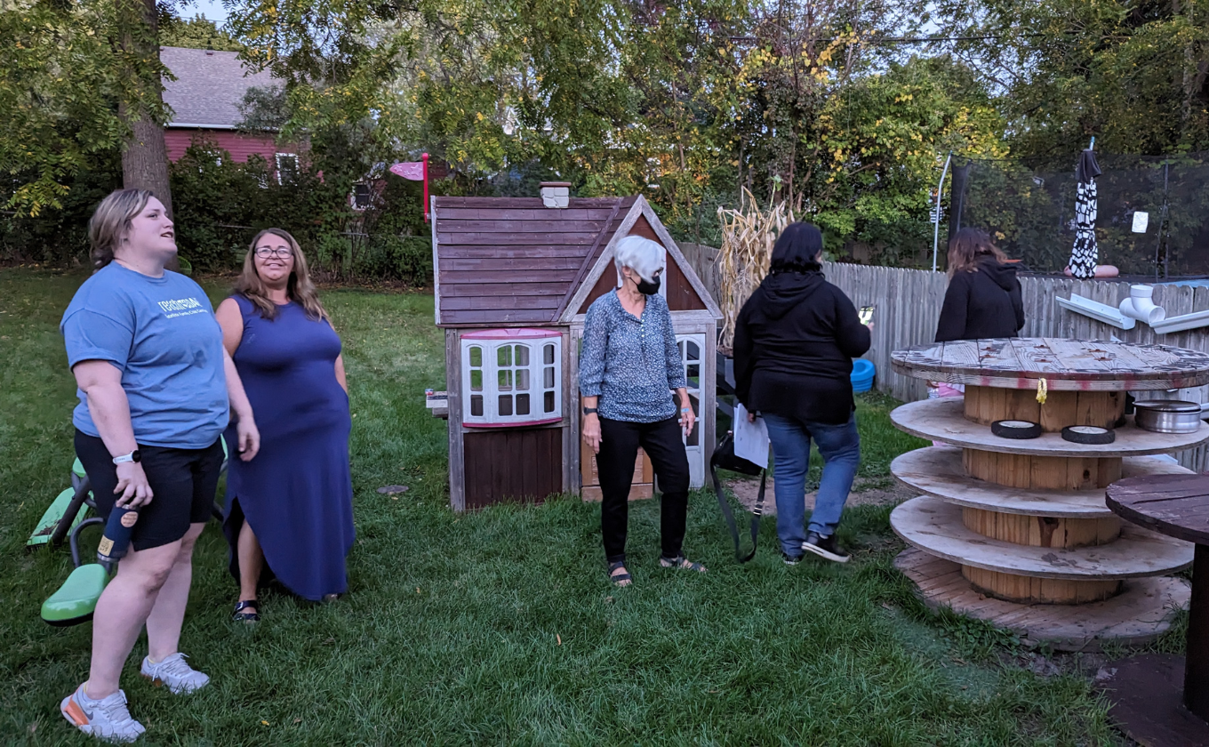 Five women standing and walking near a small playhouse and round wooden tables in a backyard.