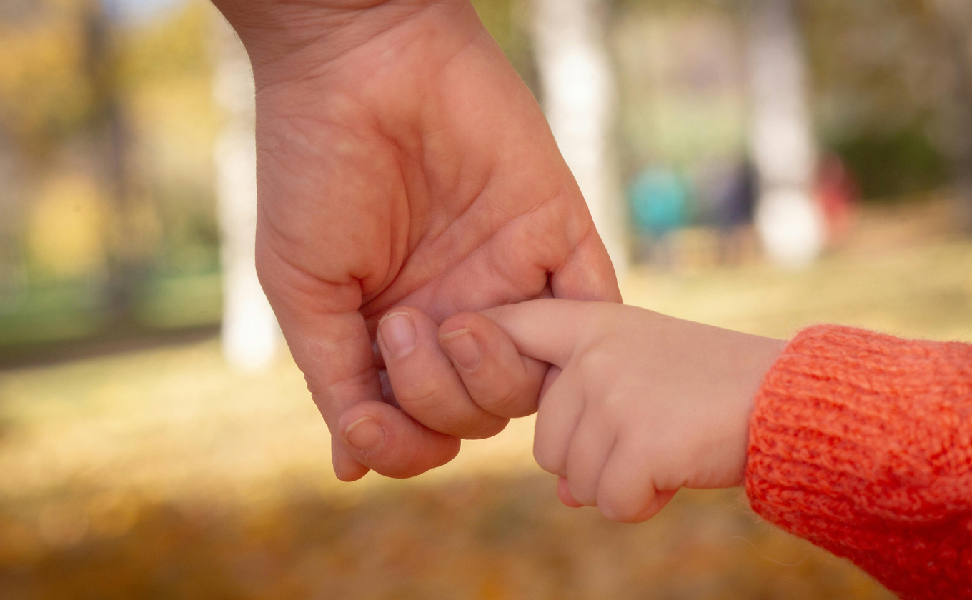 Close-up of adult and child holding hands with blurred outdoor background.
