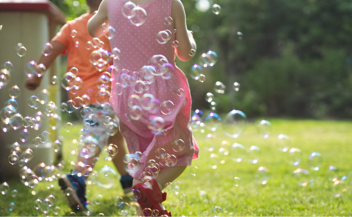 Two children running outdoors through many floating soap bubbles on a sunny day.