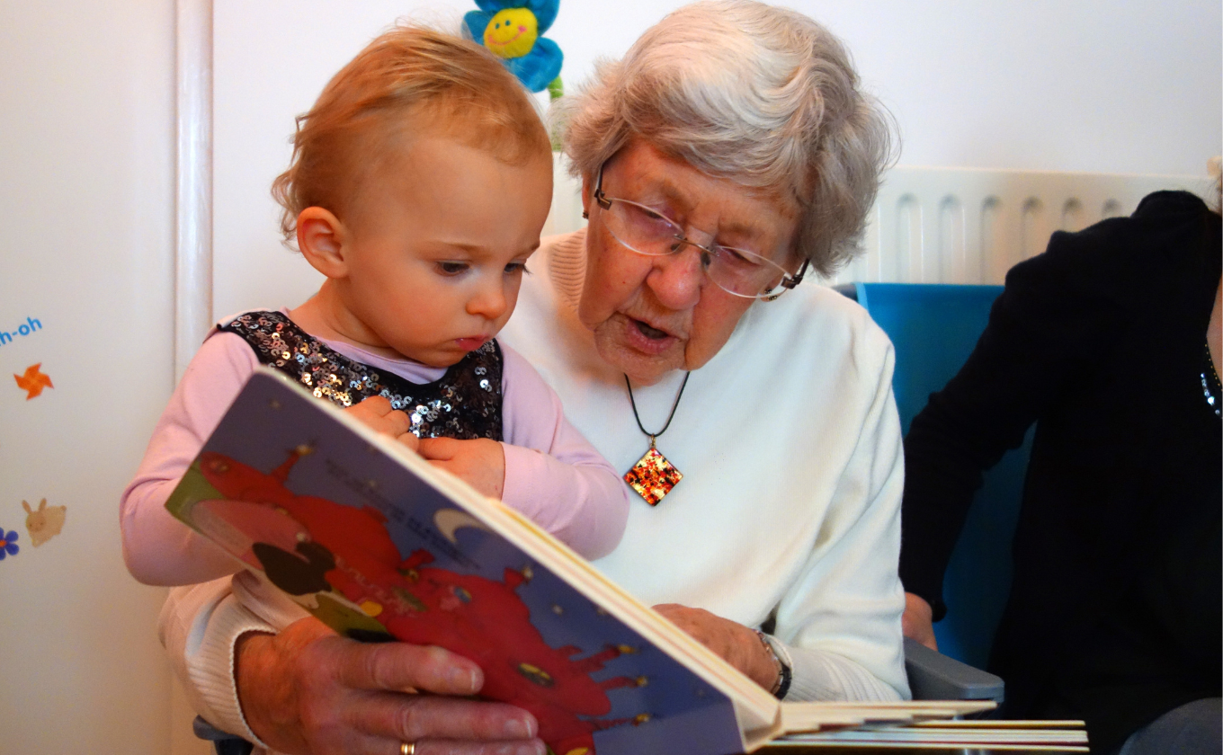 Older adult reading a colorful book to a focused young child sitting on her lap.