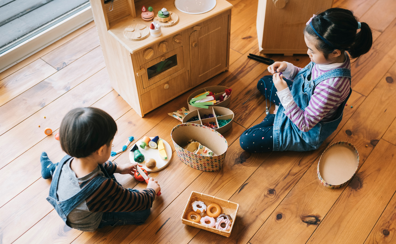 Two children sitting on wooden floor playing with toy kitchen and pretend food items.