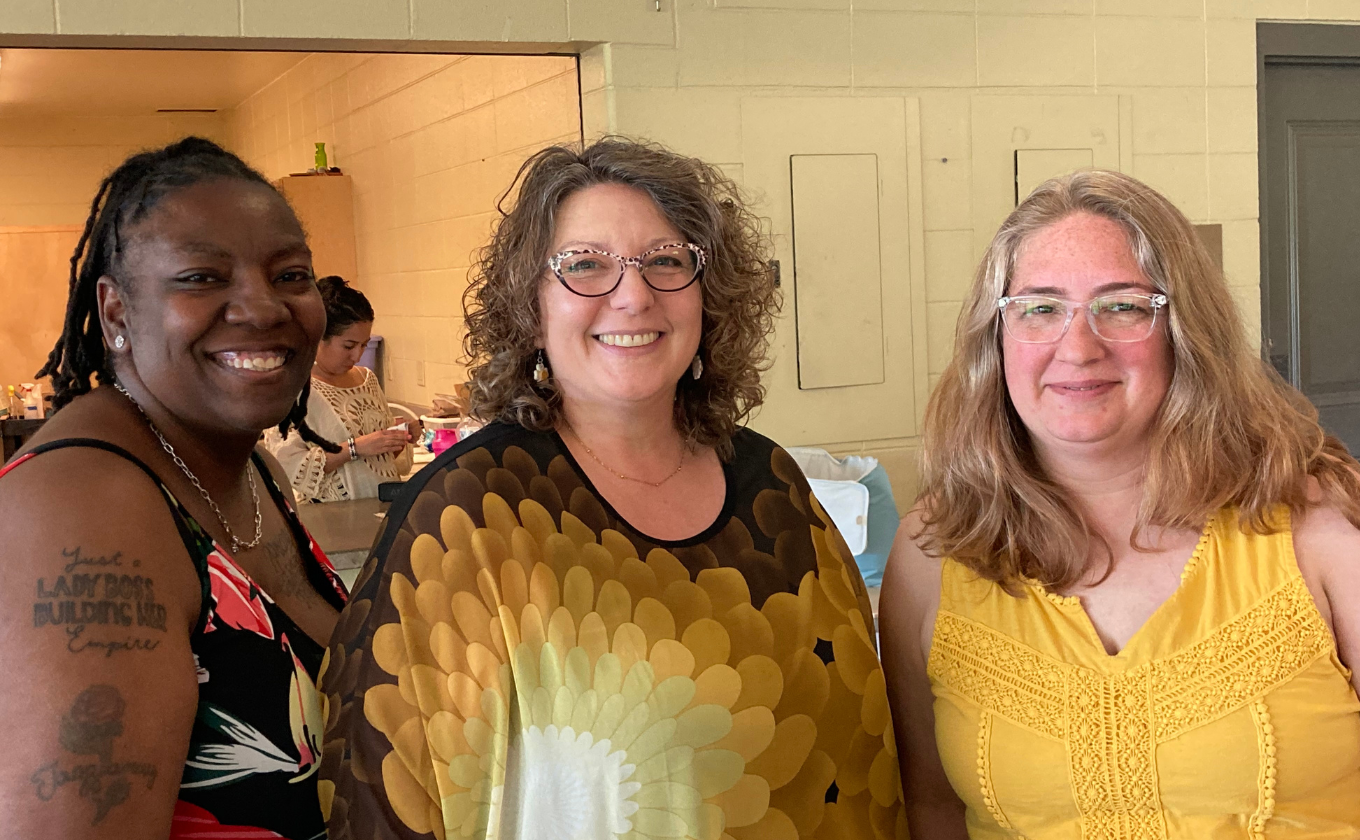 Three smiling women standing indoors, one with glasses and curly hair, one with tattoo and dreadlocks.
