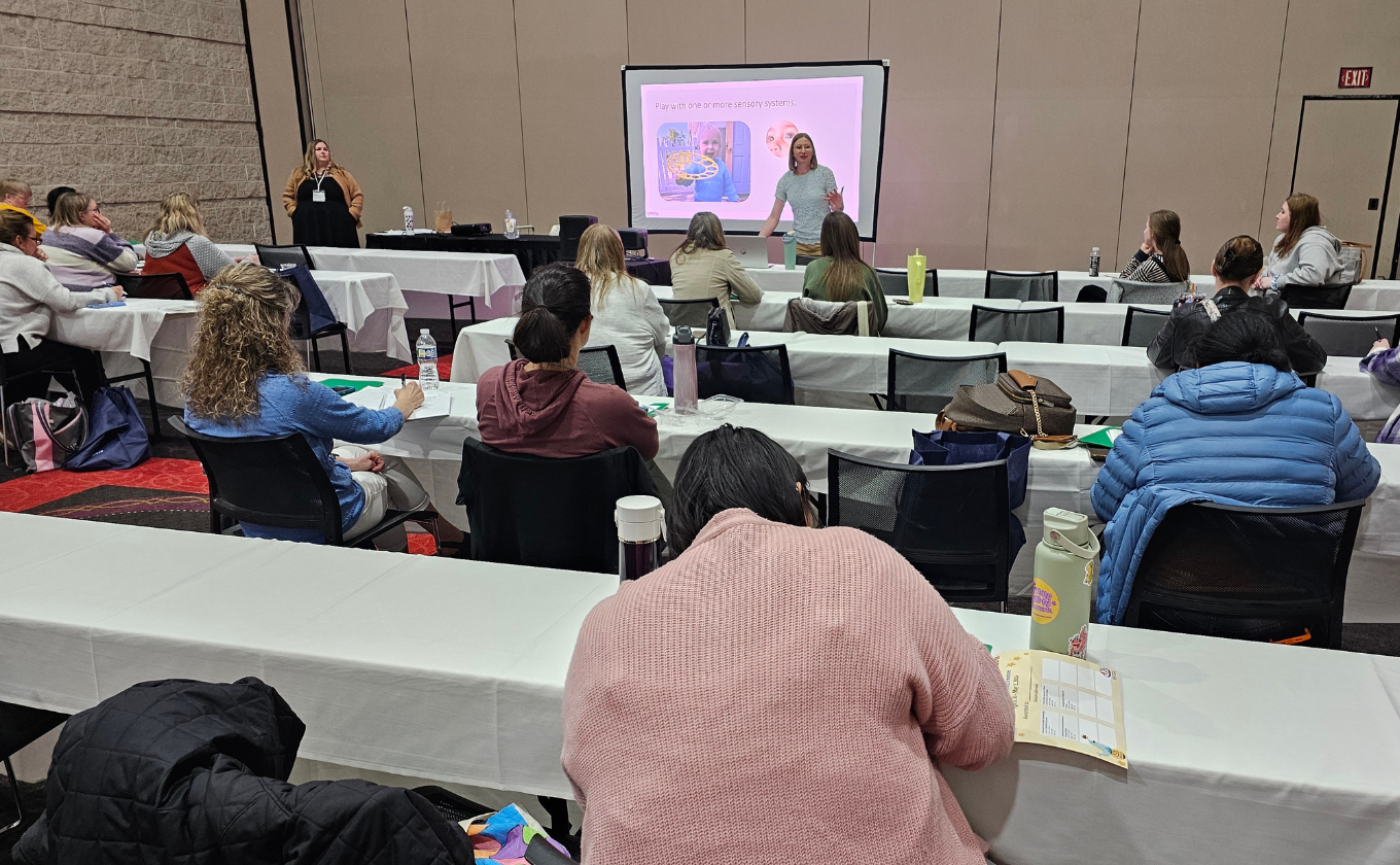 Instructor presenting to a seated audience in a conference room with rows of tables and chairs.