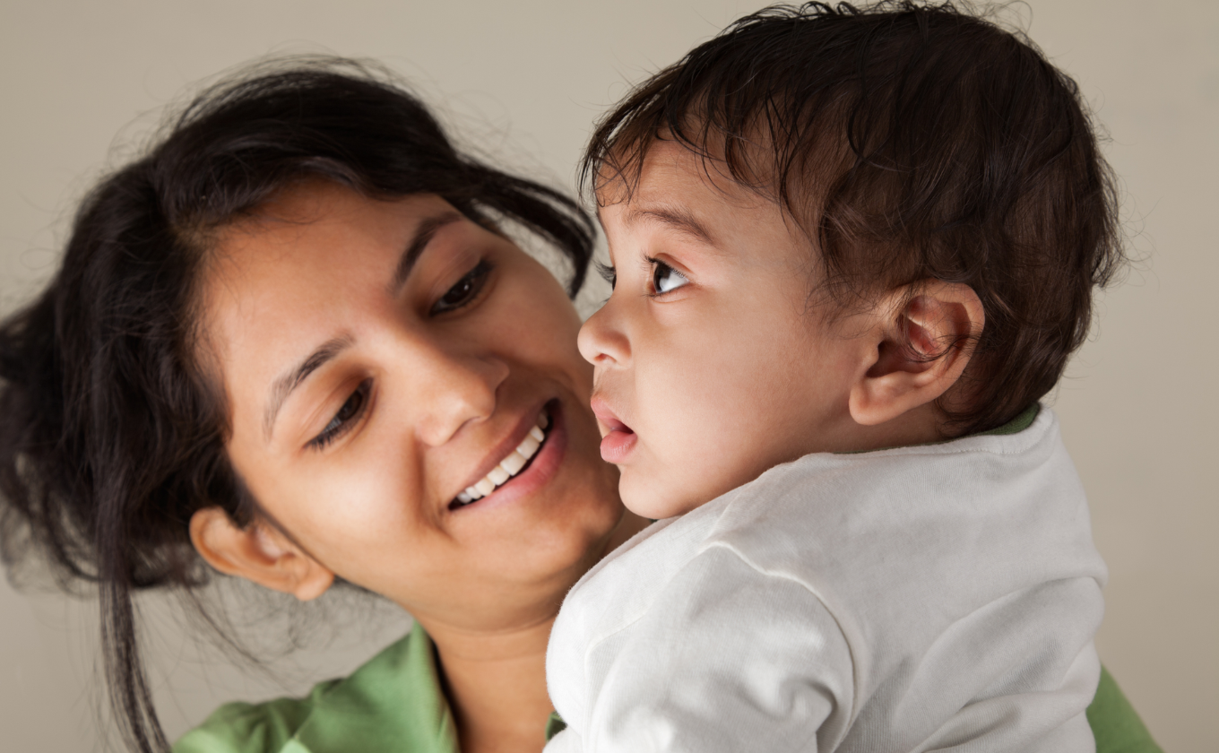 Smiling woman holding a young child dressed in white against a neutral background.