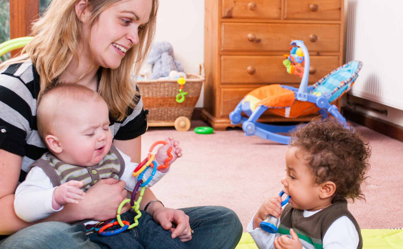 Woman holding baby with toy rings while another baby chews a toy in a cozy room.
