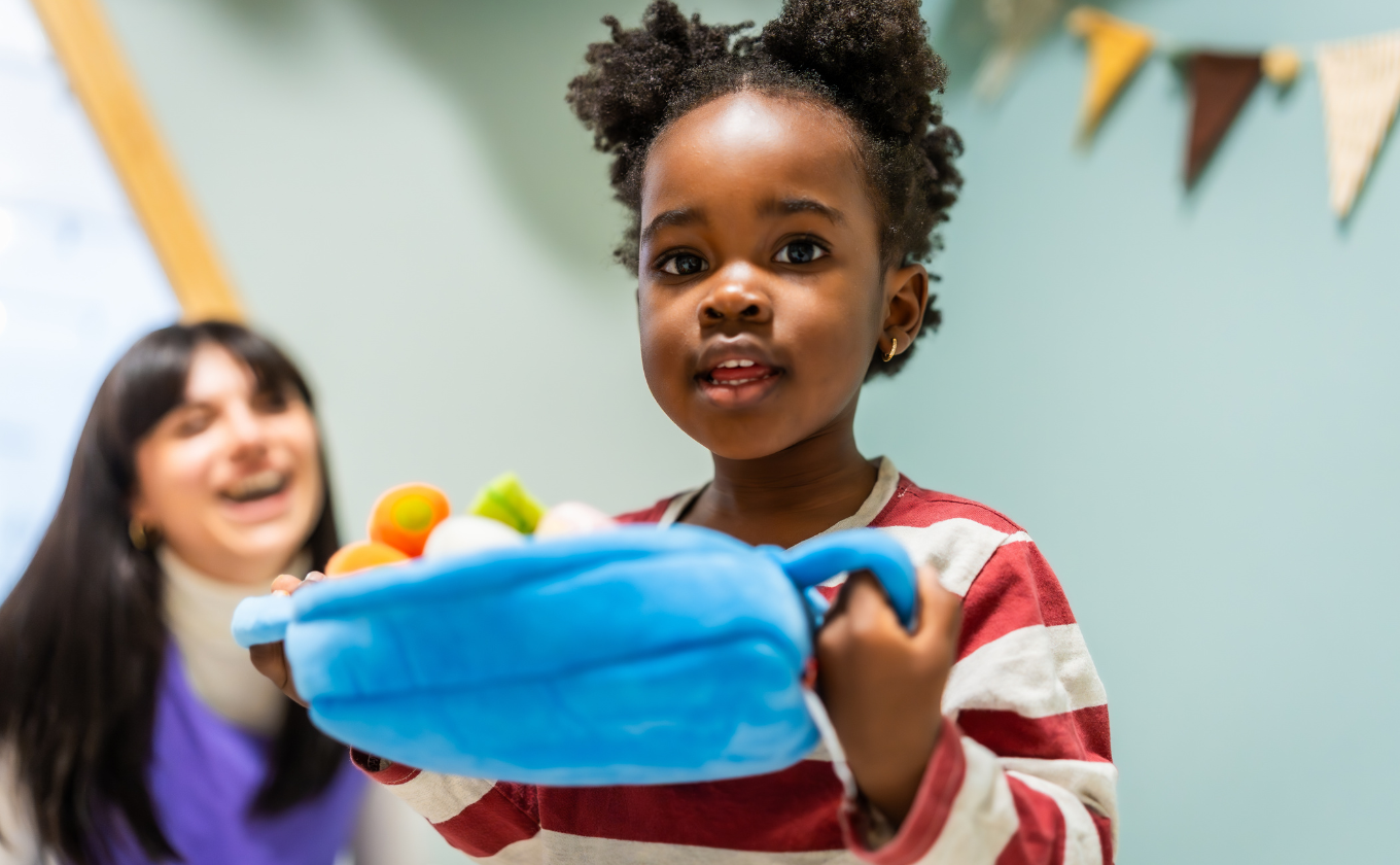Child holding a blue plush basket with toy fruits, woman smiling in background.