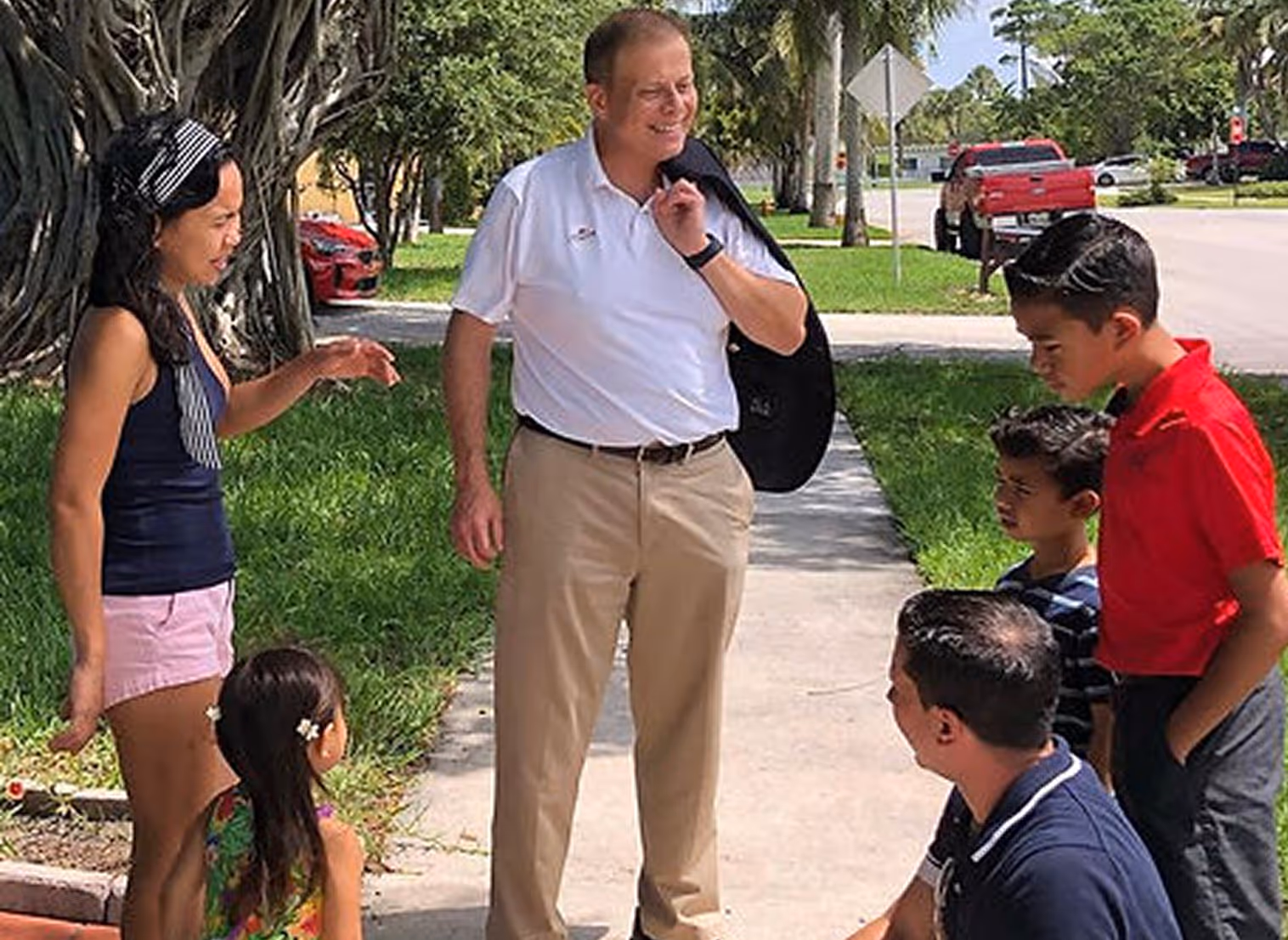 A man in a white polo shirt holding a jacket over his shoulder smiles while talking to a group of children and a woman on a sunny sidewalk.