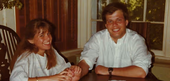Smiling young man and woman sitting at a table indoors, holding hands and looking at the camera.