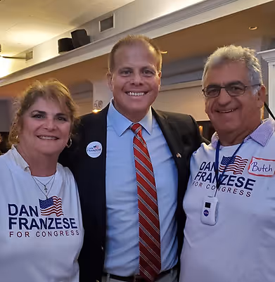 Three people smiling indoors, with the man in the center wearing a suit and red striped tie, and the two on the sides wearing white 'Dan Franzese for Congress' campaign shirts.