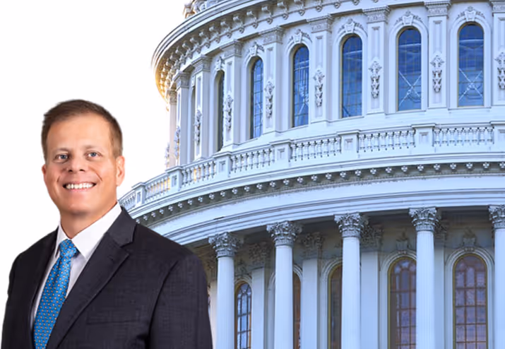Smiling man in a dark suit and blue tie standing in front of a classical government building with columns and arched windows.