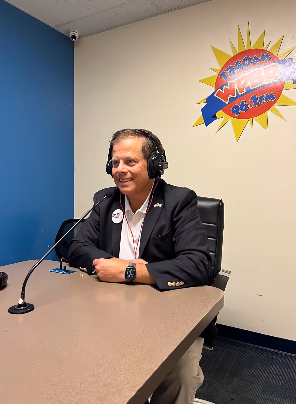Man wearing headphones and a suit jacket speaking into a microphone in a radio studio.