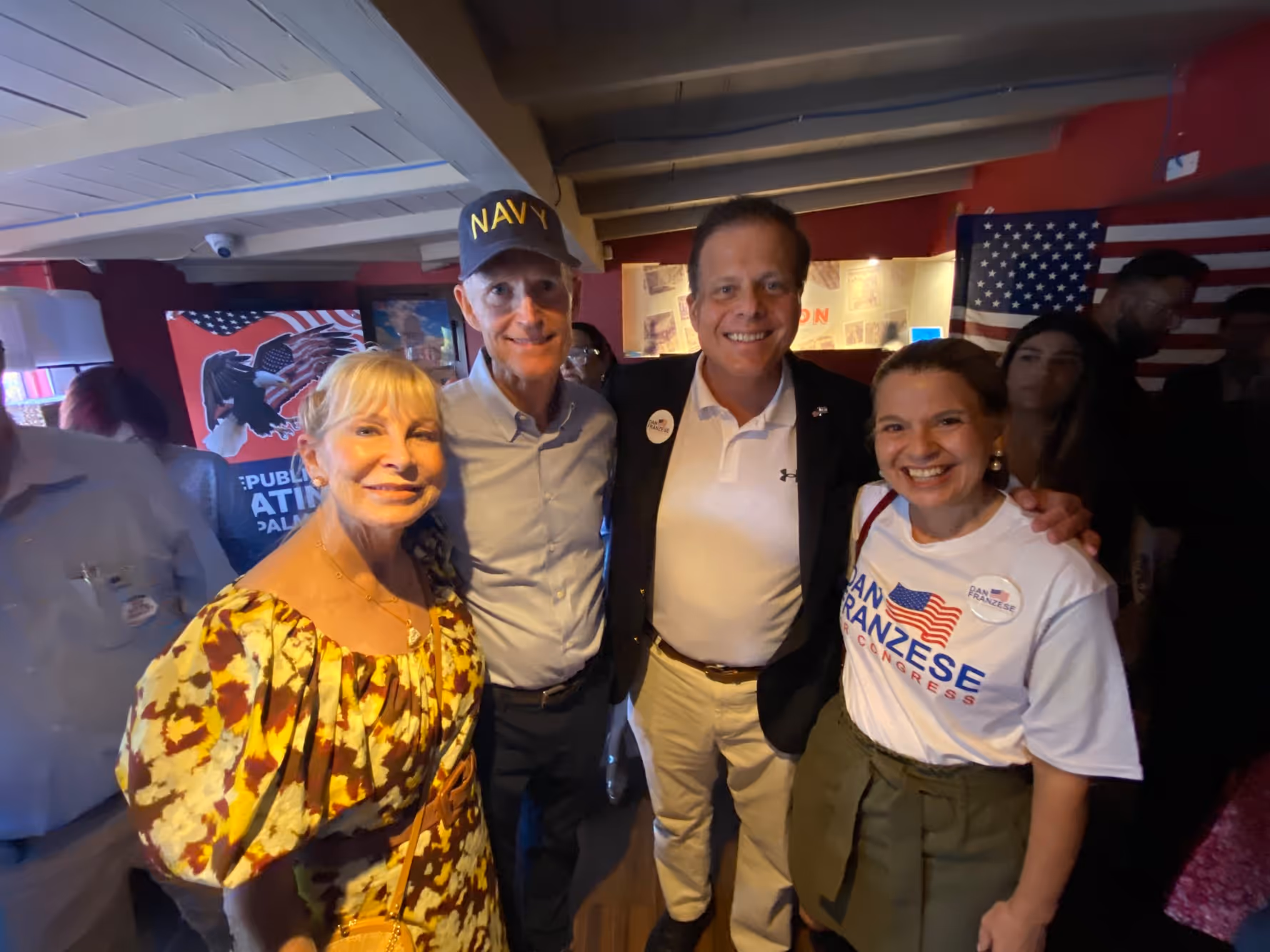 Four people smiling indoors at a political event with American flags and campaign signs.