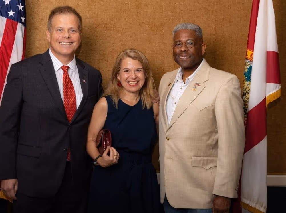 Three people standing and smiling in front of a brown wall, with American and Florida state flags in the background.
