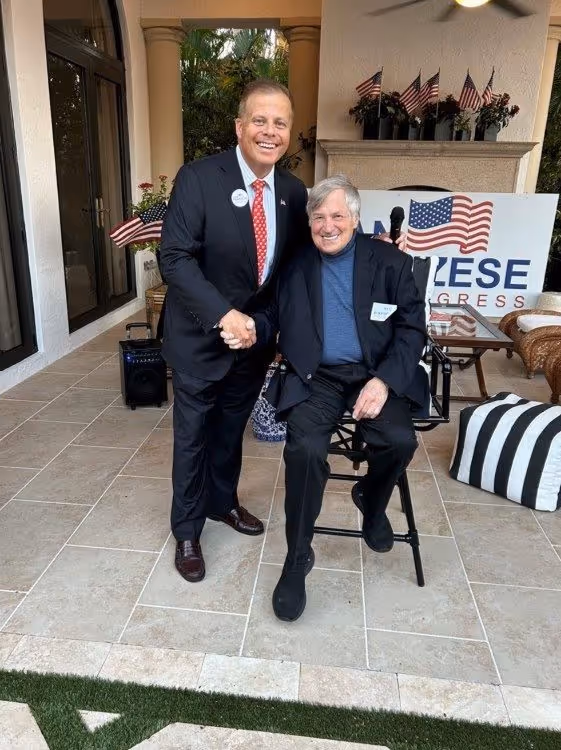 Two men smiling and shaking hands on a patio with American flags.