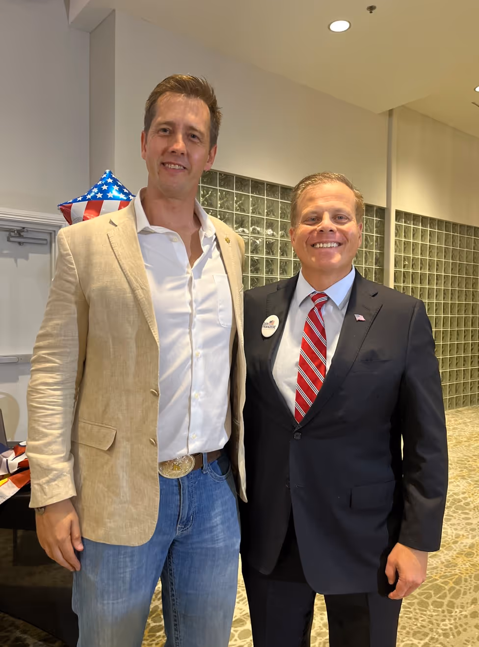 Two men smiling indoors, one in a beige blazer and jeans, the other in a dark suit with a red striped tie.