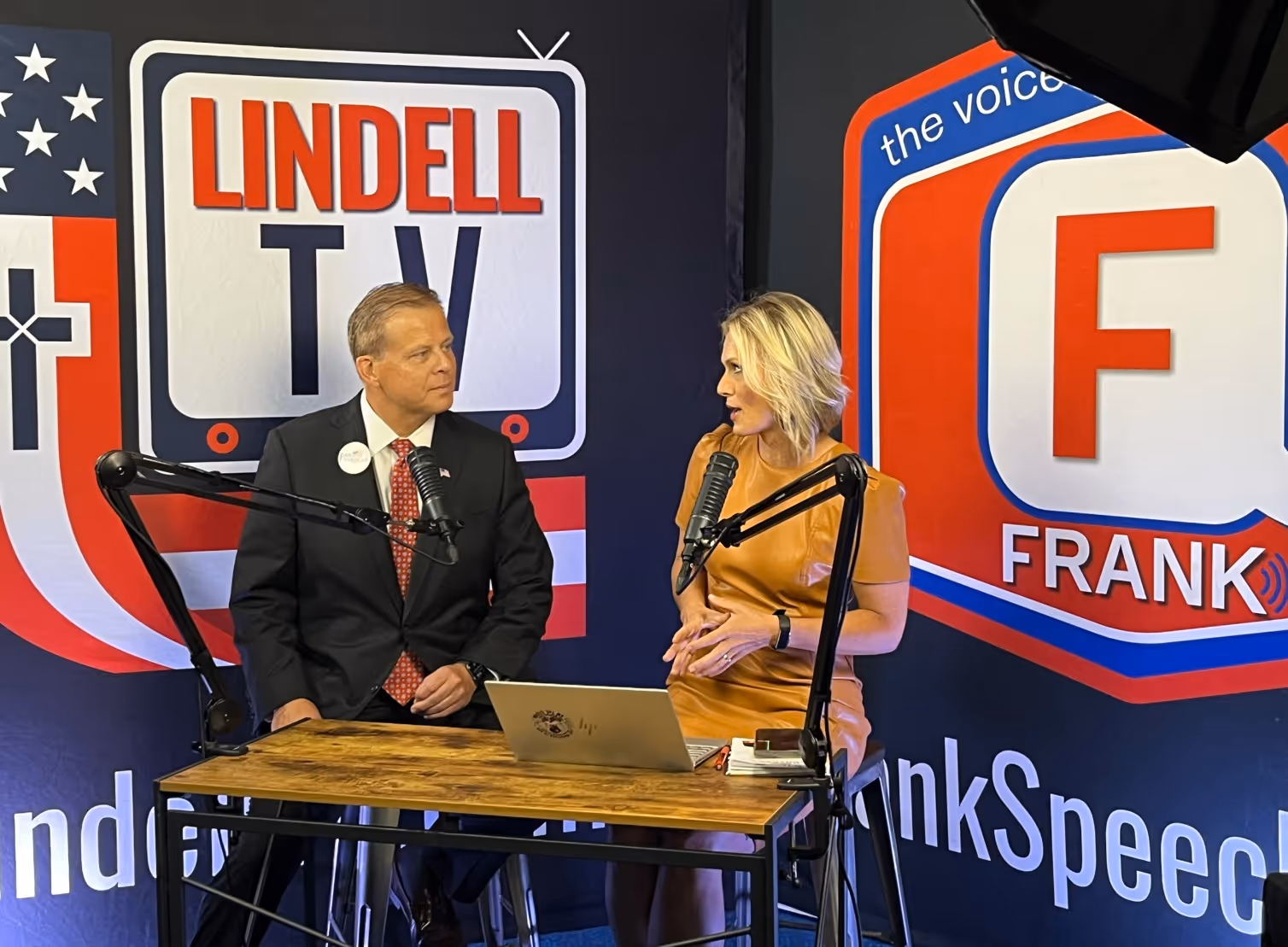 Two people sitting at a wooden table with microphones, engaged in a discussion on a set.