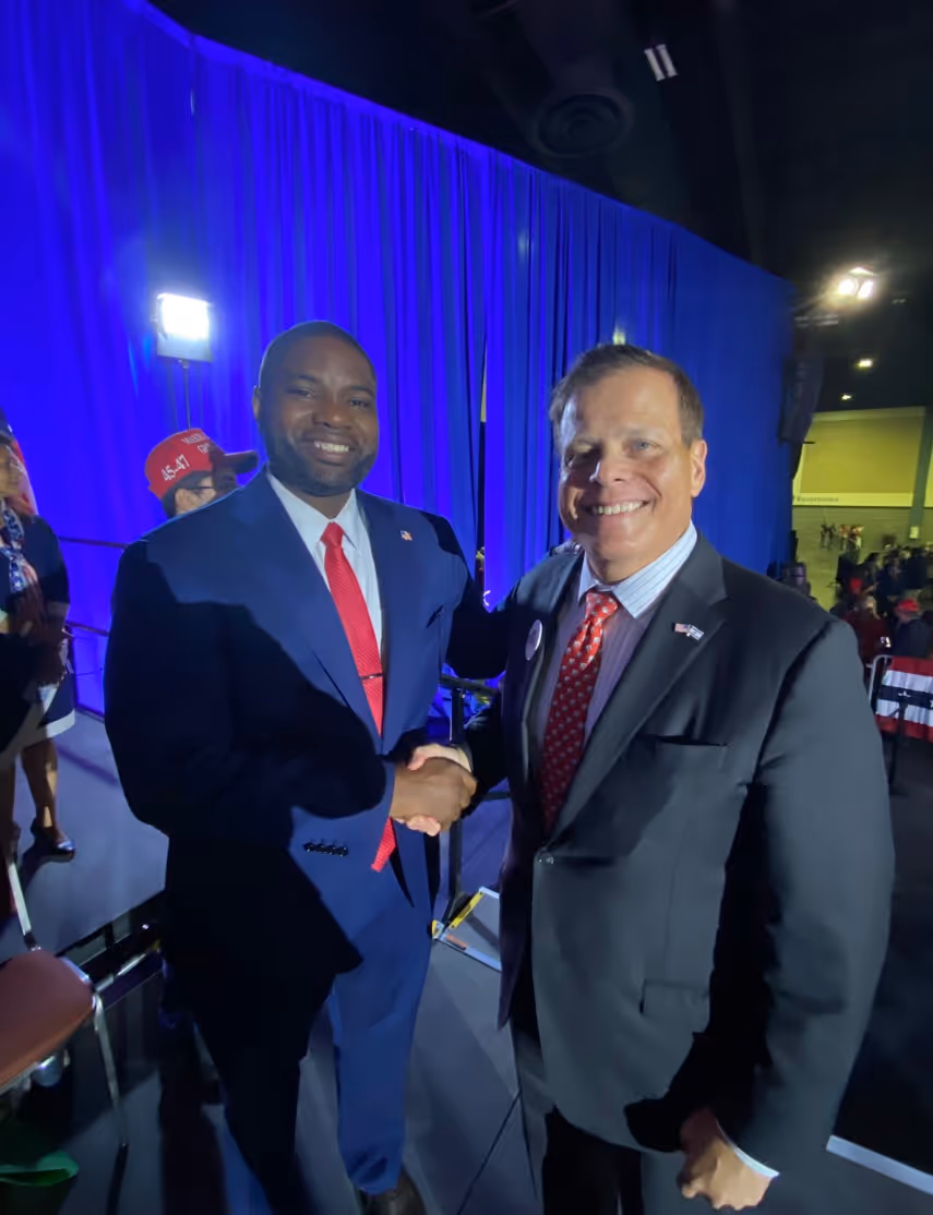 Two men in suits shaking hands, standing indoors in front of a blue curtain.