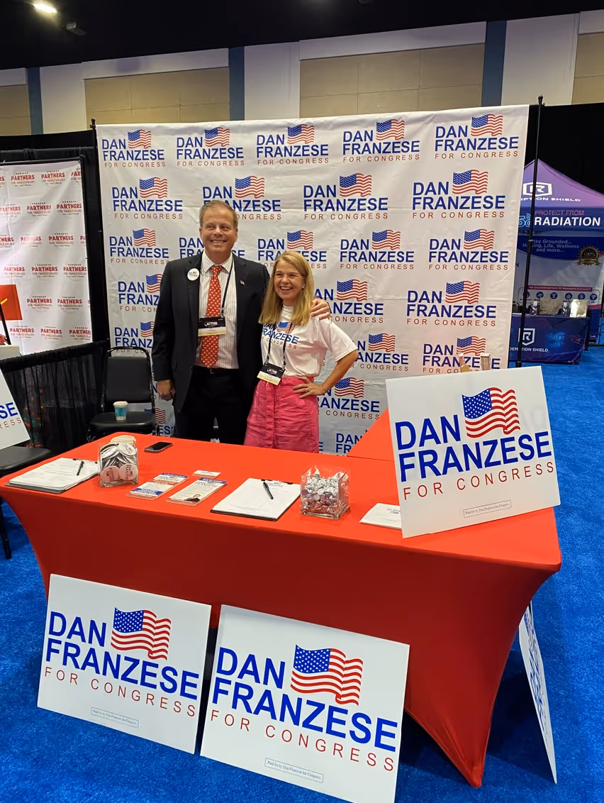 Two people standing behind a red-covered table at a Dan Franzese for Congress campaign booth with multiple signs.