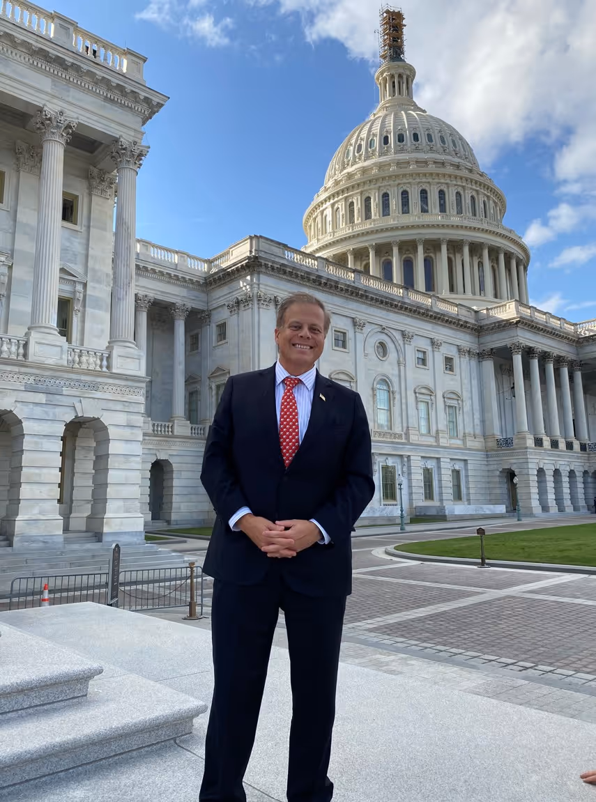 Man in a dark suit and red tie standing in front of the U.S. Capitol Building under a partly cloudy sky.