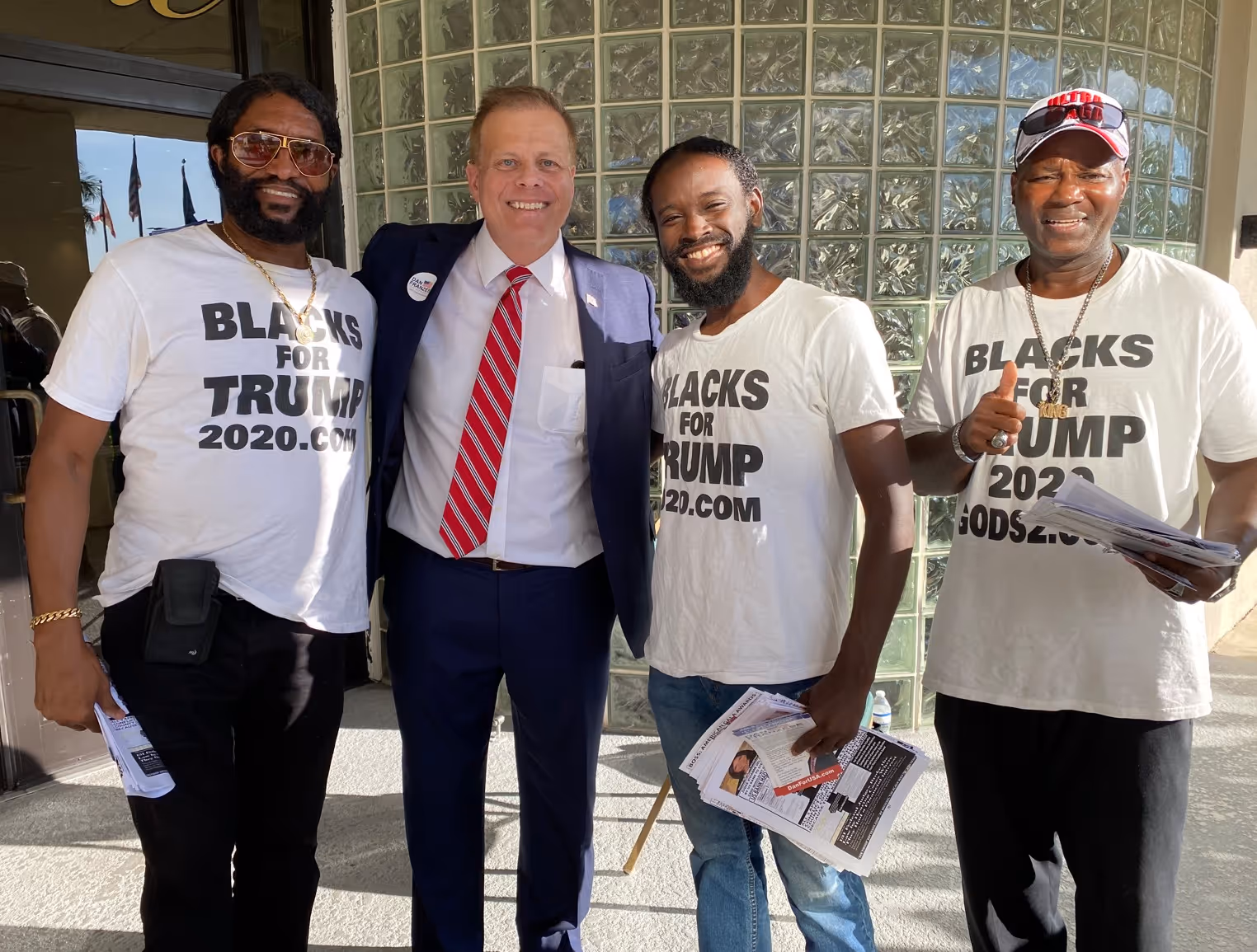 Four men standing together, three wearing white t-shirts reading 'Blacks for Trump 2020.com,' one in a suit and red striped tie.
