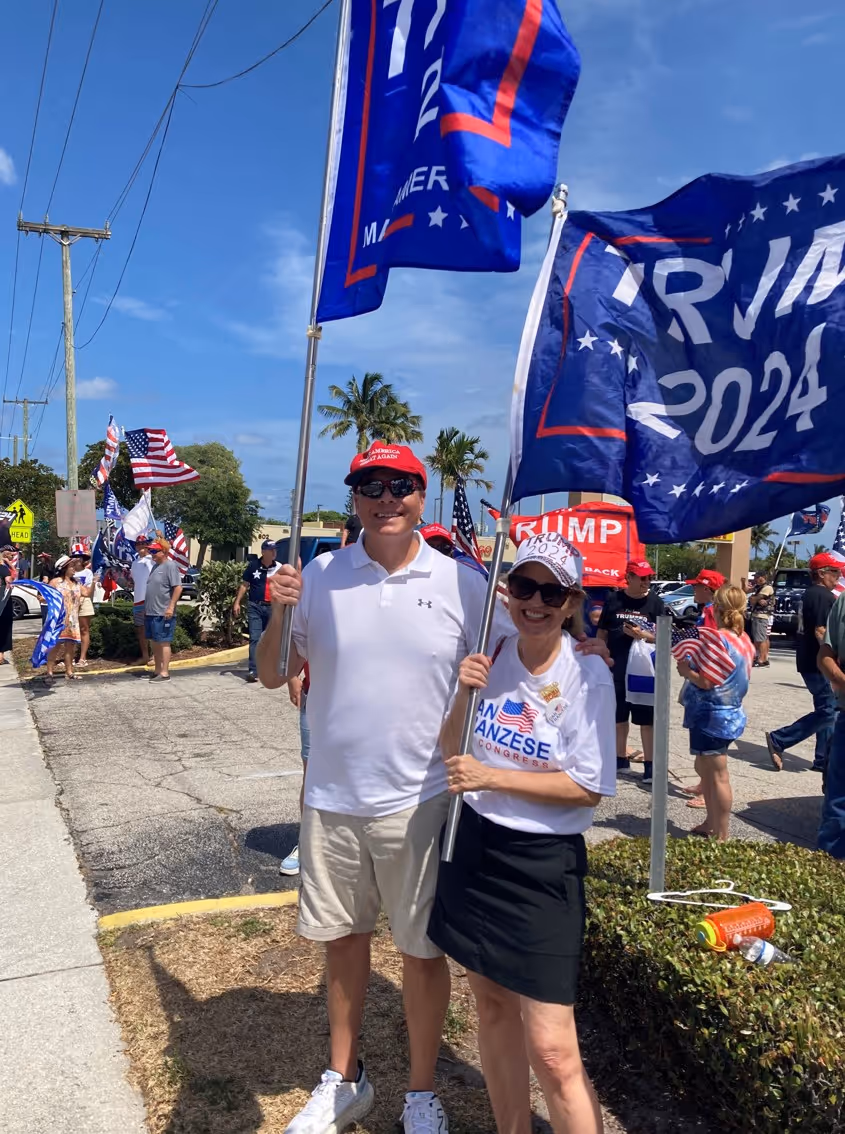 Two people smiling and holding Trump 2024 flags at a political rally.