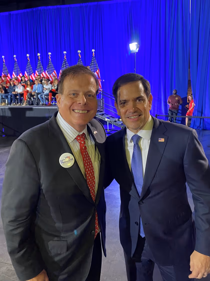 Two men in suits posing and smiling at an event with American flags.
