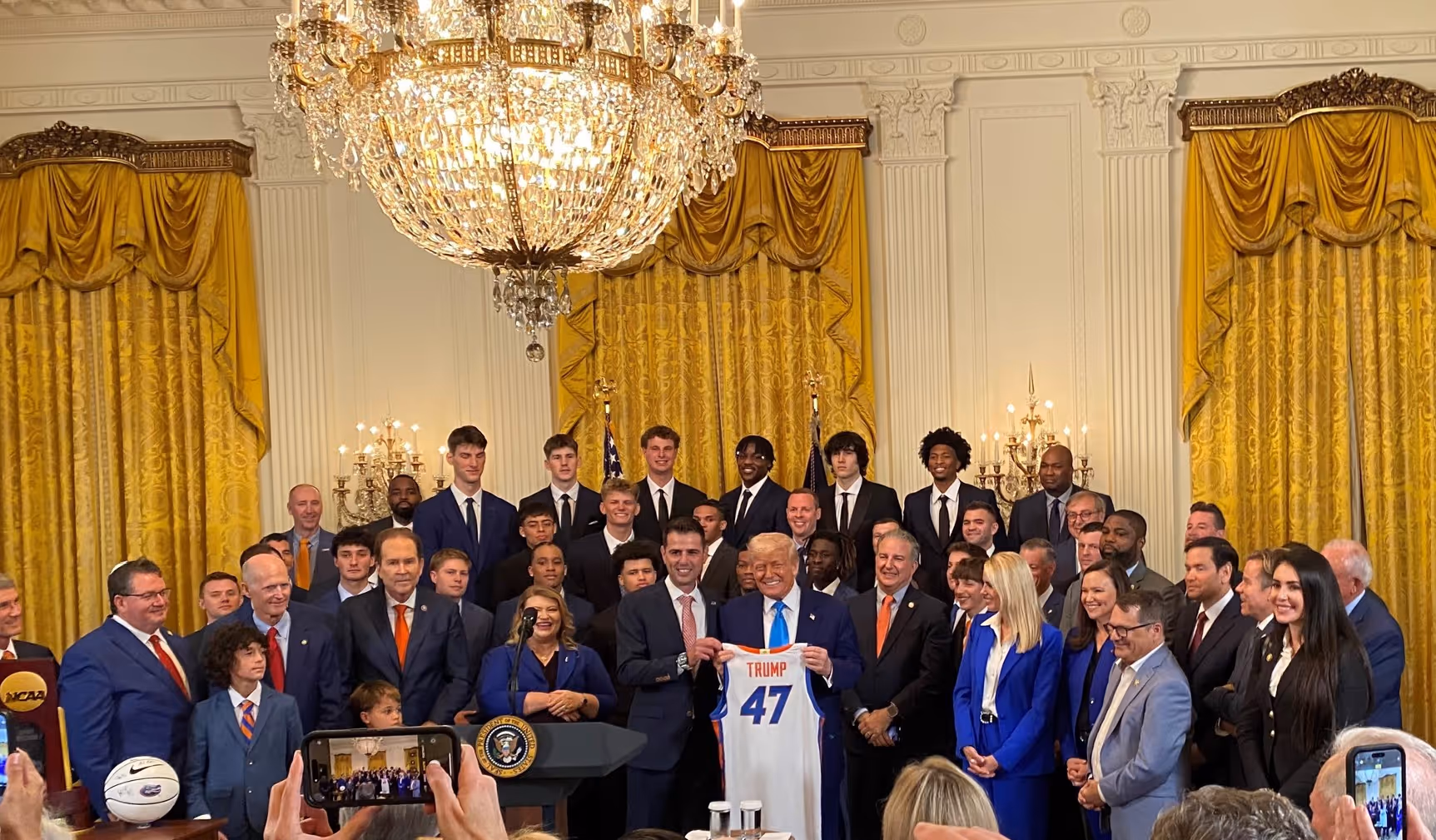 Former US President Donald Trump holding a basketball jersey with the number 47, surrounded by a large group of people.