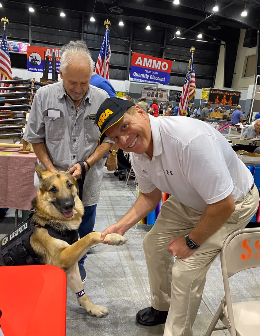 Man wearing an NRA cap smiling and shaking paws with a German Shepherd service dog at an indoor event.
