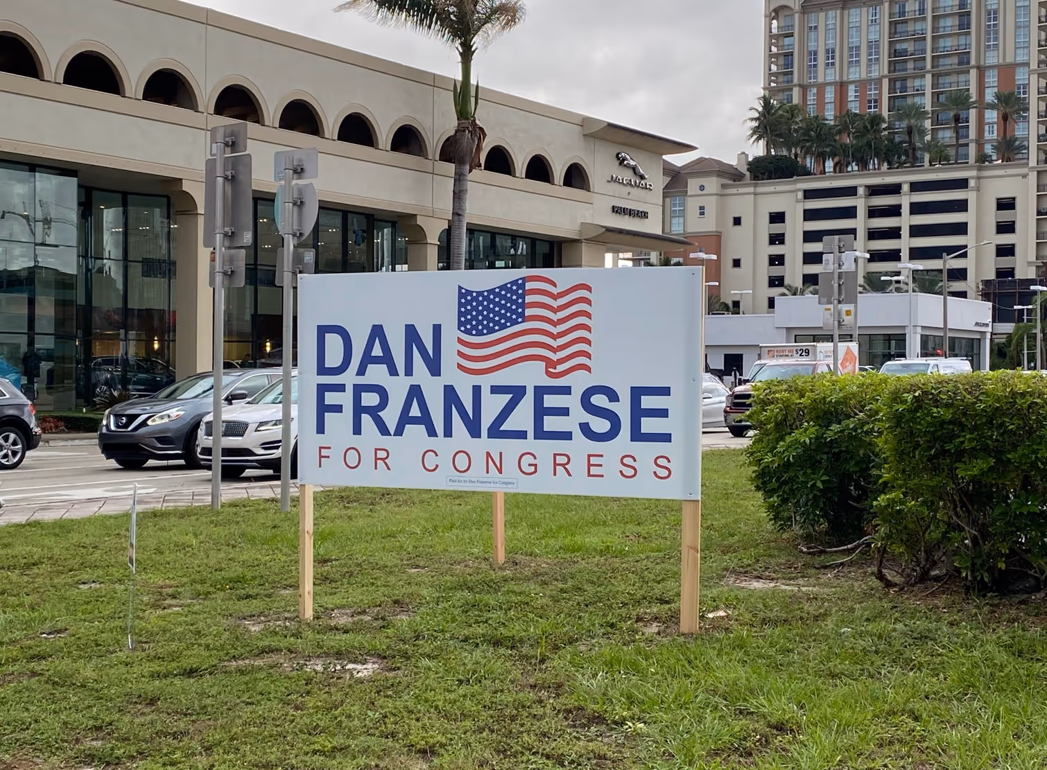 Political campaign sign for Dan Franzese for Congress with an American flag graphic on a grassy area near a street and buildings.