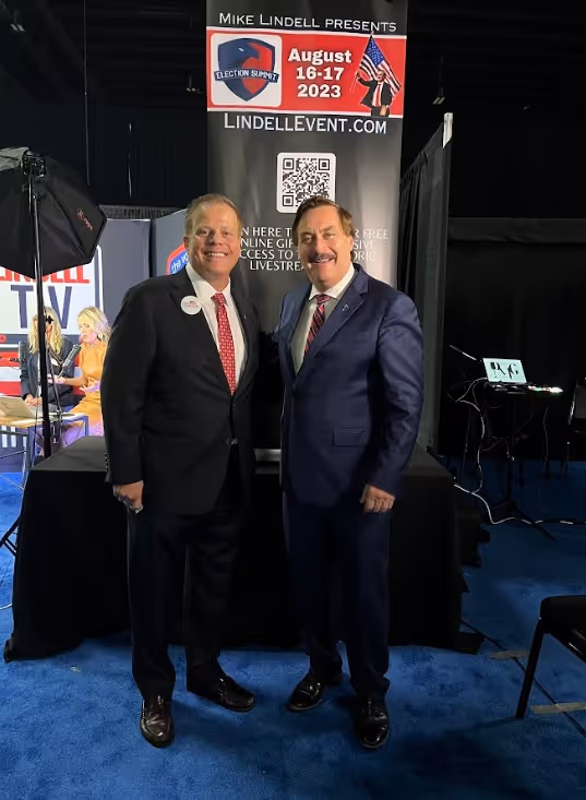 Two people smiling outdoors wearing Dan Franzese for Congress t-shirts and holding campaign flyers.