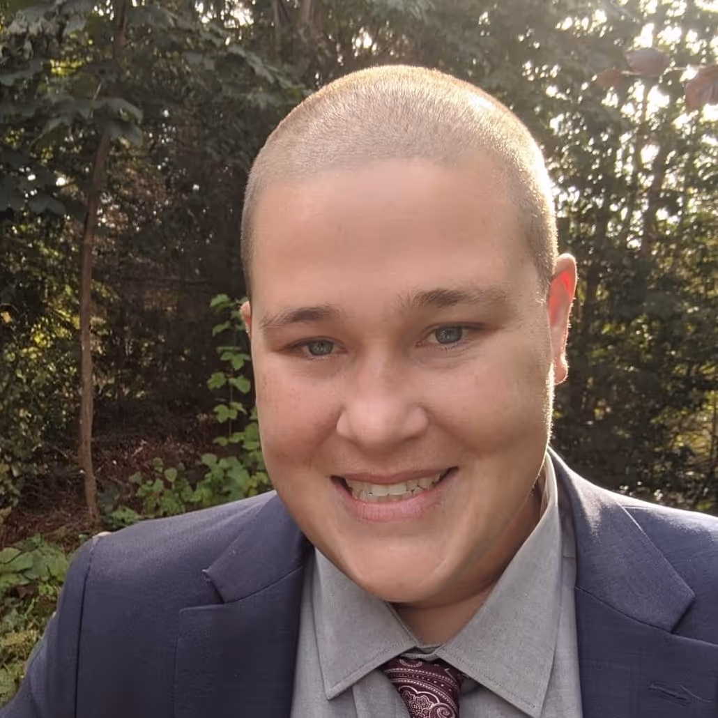 Smiling person with short hair wearing a suit, tie, and shirt outdoors in a leafy background.